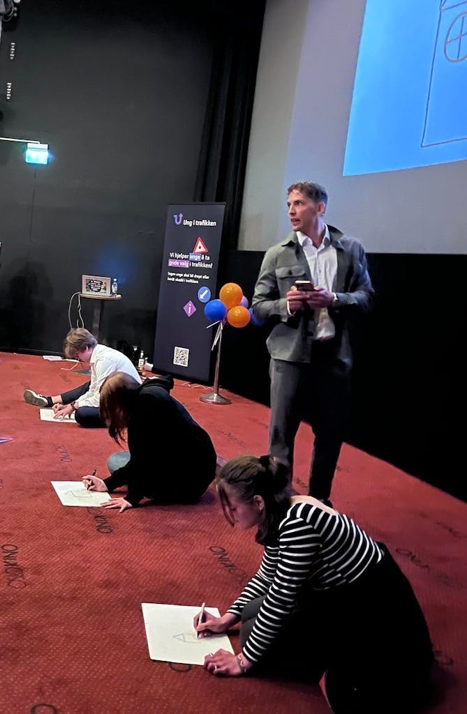 Lars Henriksen regisserer tegnekonkurranse. 3 elever sitter på gulvet og tegner ifølge instrukser av Lars. 